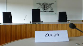 Bild zeigt den Blick in den Sitzungssaal. Im Vordergrund den Vernehmungstisch des Zeugen. Im Hintergrund die Richterbank und an der Wand das Landeswappen.